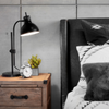 Jennifer Hughes Thumbnail Close up of light rustic wooden bedside table with a black bendable lamp and alarm clock. On right is black headboard queen bed with white stripes sheets and black furry accent pillows.