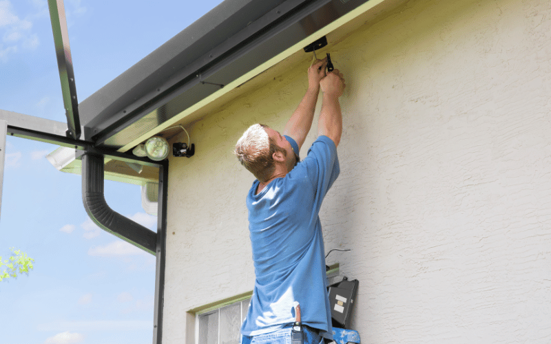 White man wearing light blue shirt on ladder mounting exterior home lighting on side of white stucco house