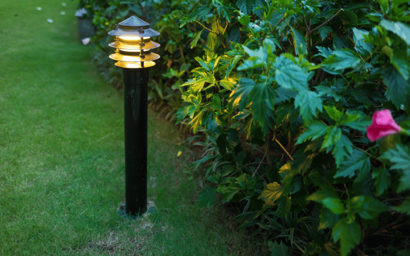 Outdoor lantern lit up on pole mounted into ground beside rose bush in dusky lighting