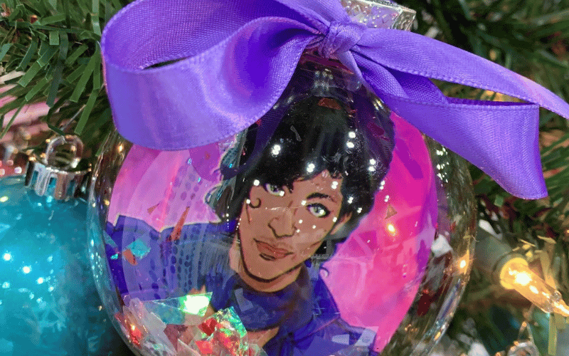 a clear Christmas ornament with a painting a man inside and a purple bow 
