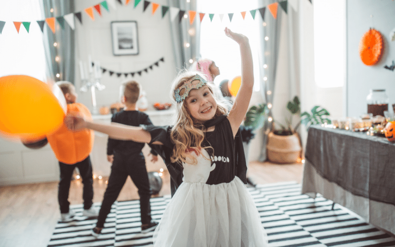 Decorated living room with Halloween pumpkins, balloons and banner. Little girl dancing wearing eye mask and ghost costume