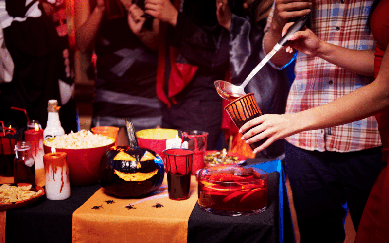 Halloween party guests at home pouring blood-like punch into a glass with Pumpkin, popcorn and Halloween decorations on a table with an orange table cloth 