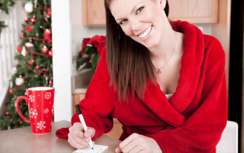 Smiling woman wearing red robe making shopping list next to red coffee cup with white snowflakes and Christmas tree in background