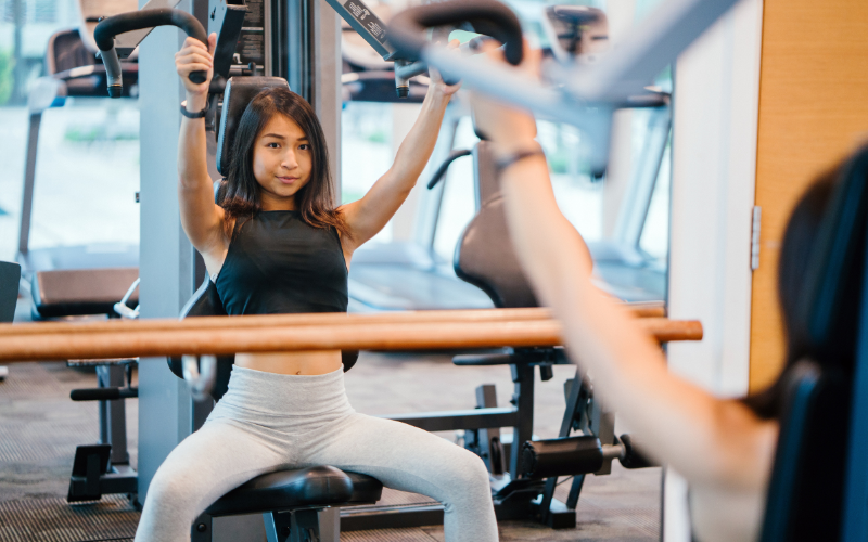 young woman with dark hair using an arm exercise machine in front of a mirror wearing a black tank top and light grey workout pants