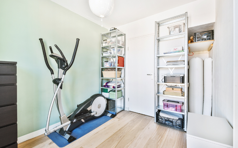 Gazelle exercise bike in storage room beside black dresser and metal storage shelves in white and light green room with light hardwood floors