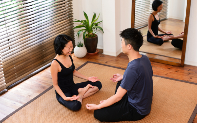 Heterosexual Asian couple sitting on brown carpet on floor doing yoga facing each other in front of mirror next to large green plant