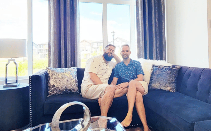 Stephen & Branden the visionary husbands sitting on blue velvet couch in front of big bight window with brown curtains