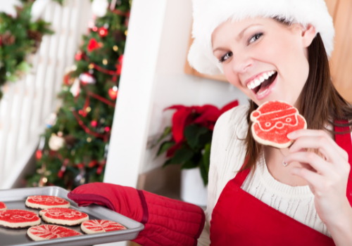 Woman wearing red apron red oven mits and Santa hat smiling and holding tray of decorated sugar cookies