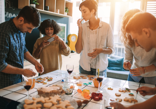 Group of young adults drinking red wine and decorating holiday cookies on a white tiled island in a bright kitchen