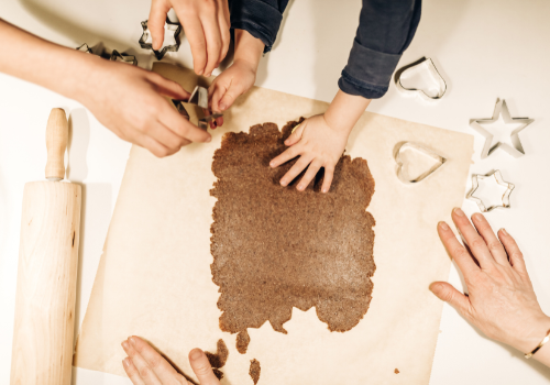 adult female hands help child use cookie cutters on flattened dough on white counter next to rolling pin