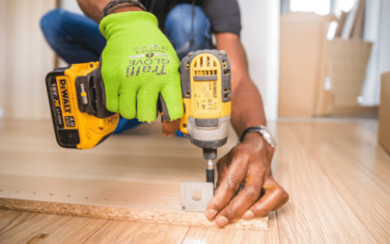 Close up of Black male professional contractor wearing neon green gloves holding yellow drill putting brackets into wooden piece