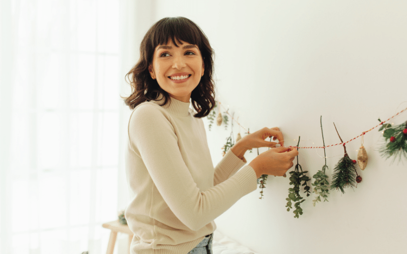 Woman with dark hair wearing off white long sleeved shirt hanging up pine decor on existing string on white wall