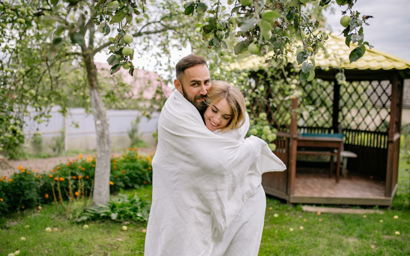 Couple standing in backyard wrapped in white duvet snuggling in front of backyard gazebo