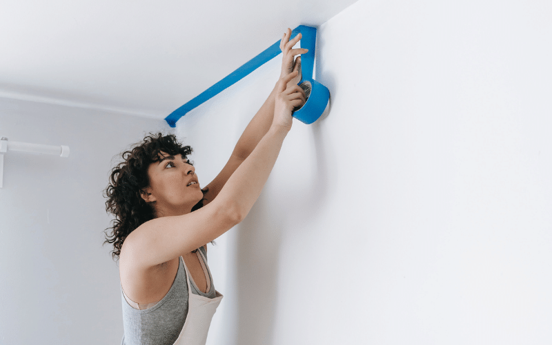 Woman with dark curly hair wearing white apron over grey tank top putting up blue painters tape on wall