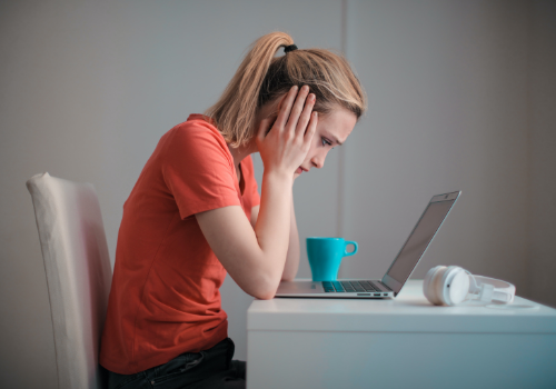 Sad girl looking at laptop with her hands on her cheeks with blue coffee cop next to laptop