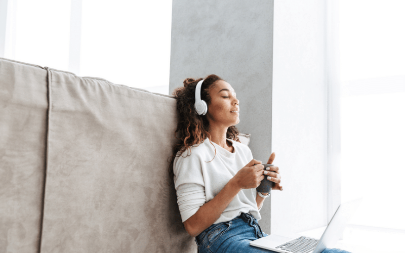 Young mixed race girl with curly hair listening to something on white over ear headphones drinking from a black coffee up with laptop on her lap leaning against grey suede couch