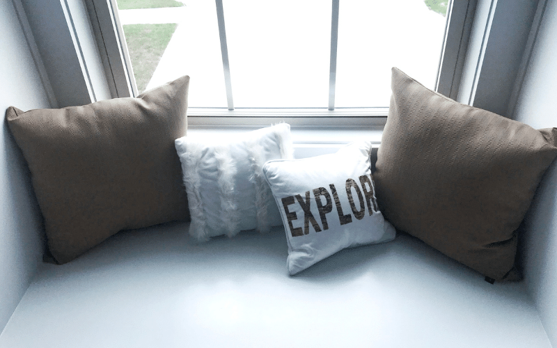 Seating area in second floor window with four pillows. Brown medium pillow then fluffy off-white pillow, white pillow with explore written on it and another brown pillow