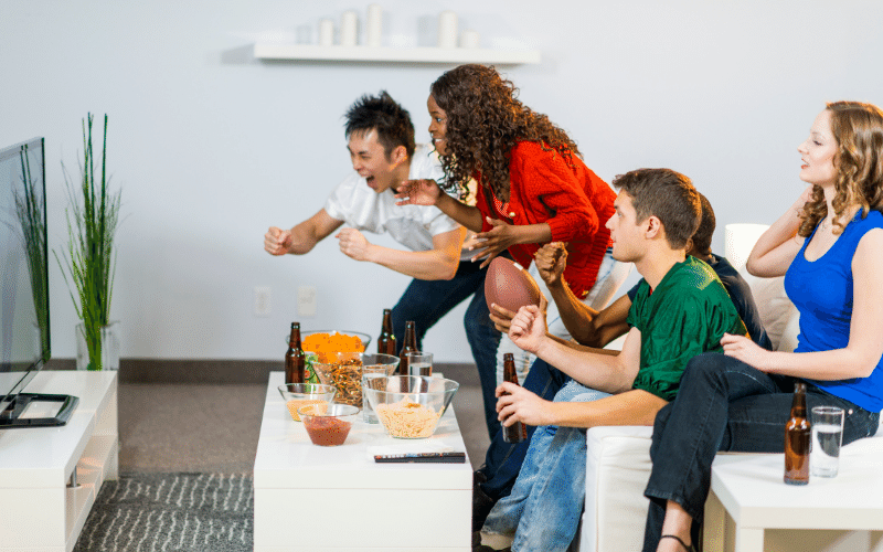 five people sitting on a white couch watching tv and cheering at the screen