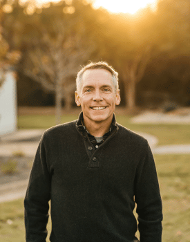 Clint Harp smiling wearing black sweater standing outside in sunlight in front of trees on grass