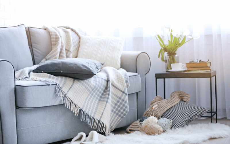 Grey couch with white plaid blanket, white shag rug, navy pillow and balls of yawn for knitting on floor in front 