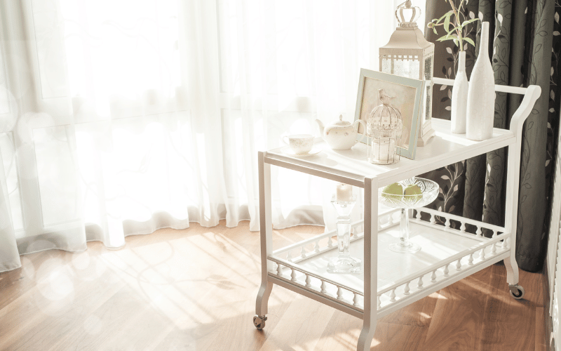 White bar cart with bottles of wine, tea pot, picture frame and fruit on it. 