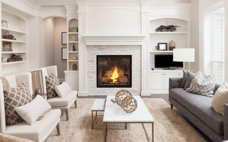 Modern white living room with beige patterned throw pillows and a medium grey couch in front of a lovely brick fireplace with gold accent decorations on the marble coffee table