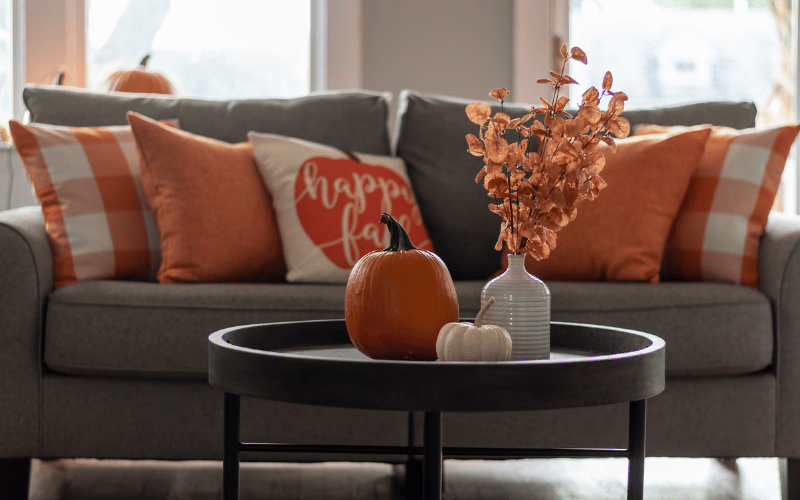 Interior design orange and white pillows plus pumpkin pillows, orange fake flowers and an orange and a white pumpkin on a table in front of a grey couch
