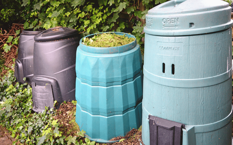 2 grey and 2 blue tall plastic compost bins located outside next to trees