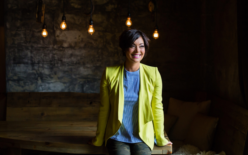 Jennifer Woch  wearing neon yellow blazer over light blue top and jeans leaning on wooden table