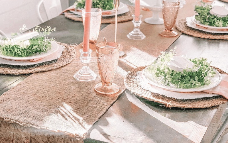 a table with a burlap runner and plates with greenery placed on it for decoration